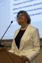 Uśmiechnięta seniorka w białej marynarce recytuje wiersz przy mównicy