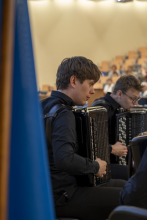 Profil młodego muzyka w czarnej koszuli grającego na akordeonie podczas występu; w tle widoczny drugi akordeonista oraz publiczność w auli.