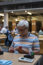 Portret uczestnika turnieju. Mężczyzna siedzi przy drewnianym stole w bibliotece i z pełnym skupieniem analizuje wachlarz kart trzymanych w dłoniach. Na stole przed nim leży białe urządzenie do zliczania punktów oraz pudełko z kartami licytacyjnymi. W tle widoczne są rozmyte regały z książkami oraz inni uczestnicy turnieju.