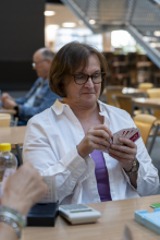 Uczestniczka turnieju z łagodnym uśmiechem układa w dłoniach trzymane karty brydżowe. Siedzi przy jasnym stole w bibliotece, w tle widoczna jest rozmyta sylwetka innego gracza.