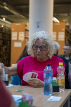 Uczestniczka turnieju siedzi przy stole w bibliotece i z uwagą przygląda się swoim kartom brydżowym. Na stole przed nią stoi pojemnik licytacyjny.