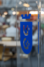 Zbliżenie na szklaną taflę, na której naklejone jest niebieskie logo Uniwersytetu Gdańskiego (UG) przedstawiające tarczę herbową z koroną i stylizowanymi literami. Tło za szybą jest mocno rozmyte, ukazując ciepłe światła i zarysy osób uczestniczących w turnieju brydżowym.