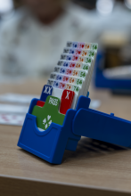 Bliskie ujęcie niebieskiego, plastikowego pojemnika licytacyjnego stojącego na jasnym, drewnianym stole. W pojemniku uporządkowane są pionowo białe karty licytacyjne z symbolami kolorów oraz plastikowe plakietki z napisami „PASS” i „X”. W rozmytym tle widać zarys postaci gracza.