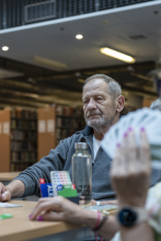 Portret uczestnika turnieju. Na pierwszym planie widać rozmyty zarys dłoni kobiety, która trzyma wachlarz kart. Na stole pomiędzy nimi stoi pojemnik licytacyjny.